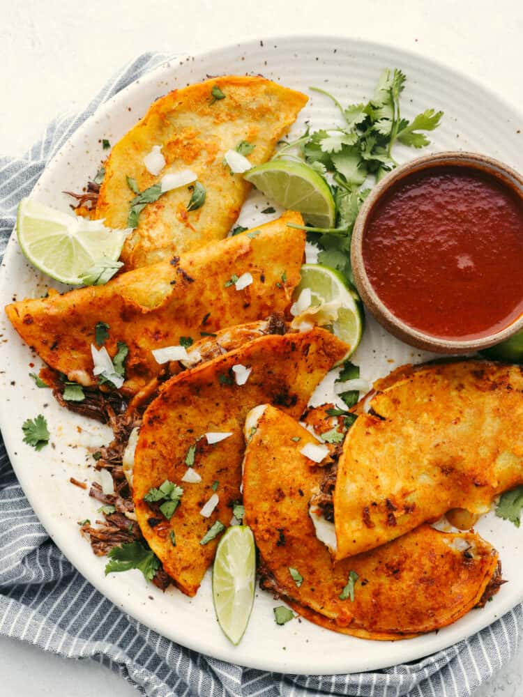 These Birria tacos are packed full of flavor! They are filled with beef and Mexican flavorings that are so delicious that you will definitely need to come to this meal hungry because you will for sure be asking for seconds! A platter with cooked birria tacos and a small bowl of sauce to dip them in. The plate it garnished with cilantro and lime slices.