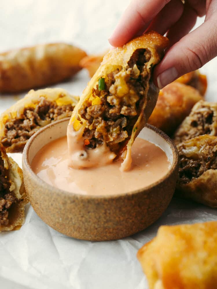 Cheeseburger eggrolls have all the beefy, cheesy goodness of a burger but inside a crispy shell! Make this delicious appetizer for your next get-together and I assure you, it will be a huge hit! Dipping a cheeseburger eggroll in a creamy sauce.