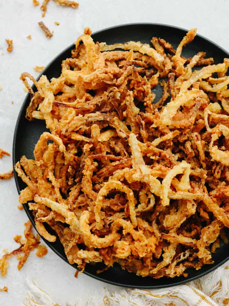 These crispy onion strings are the absolute BEST! The brown and crispy outside and the deliciously moist onion inside make this the perfect topping for any burger or salad! A black plate with cripsy onion strings.