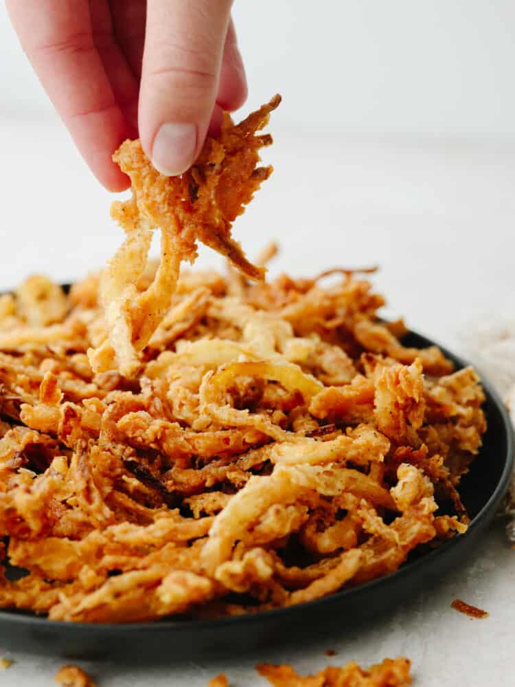 These crispy onion strings are the absolute BEST! The brown and crispy outside and the deliciously moist onion inside make this the perfect topping for any burger or salad! A hand picking up some onon strings from a black plate.