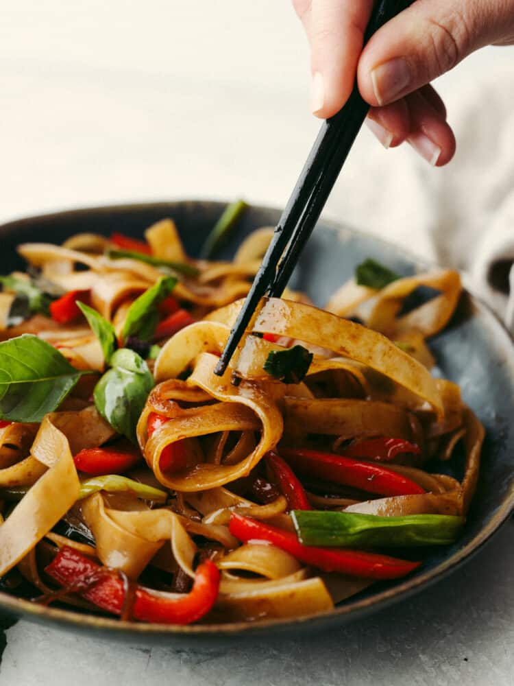 These drunken noodles (or spicy Thai noodles) are filled with delicious flavors! Coated in a perfectly savory sauce, these noodles will satisfy any Thai food craving! Drunken noodles in a bowl beaing grabbed with some black chopsticks.