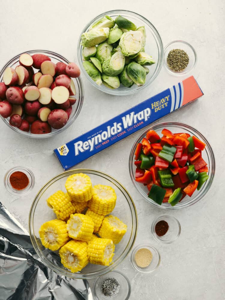 These foil pack grilled vegetables made with Reynolds Wrap® Heavy Foil are perfect for BBQ season! They cook just right on your grill and come out tender and seasoned to perfection. This is one side dish that you don't want to miss! Glass bowls of corn, peppers, potatos and brussel sprouts with a box of Reynolds foil in the middle.