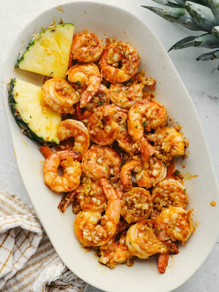 Hawaiian-style garlic shrimp is cooked up in a dreamy garlic butter sauce. Inspired by the food trucks in Hawaii, the punch of savory flavor in each bite will have you feeling like you're on vacation! Top-down view of shrimp served with pineapple in a white plate.