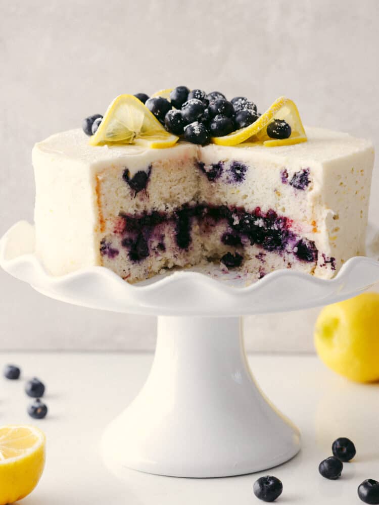 You have never seen a beautiful and equally delicious cake as this lemon blueberry cake! Elegant, delicious, and fit for any occasion, this cake will knock your socks off! A lemon blueberry cake on a white platter with a quarter of it cut out so that you can see the inside.