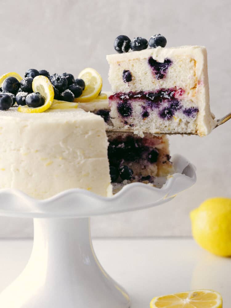 You have never seen a beautiful and equally delicious cake as this lemon blueberry cake! Elegant, delicious, and fit for any occasion, this cake will knock your socks off! The side view of the lemon blueberry cake on a platter with a slice being taken out.