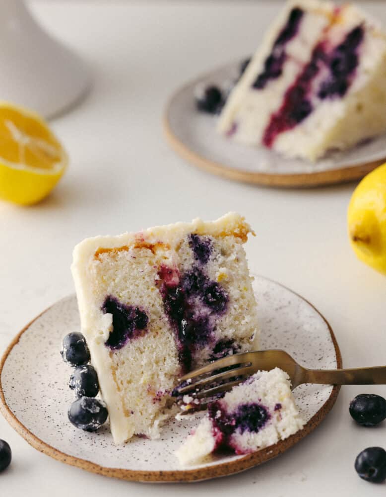 You have never seen a beautiful and equally delicious cake as this lemon blueberry cake! Elegant, delicious, and fit for any occasion, this cake will knock your socks off! A slice of lemon blueberry cake on a plate with a fork cutting into it.