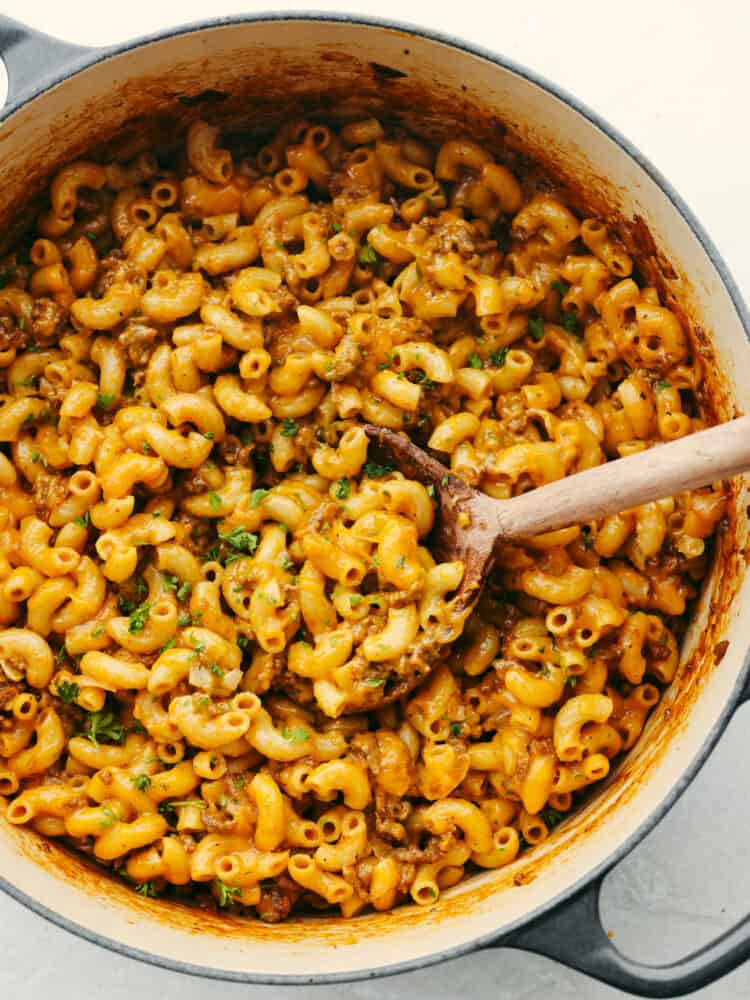 A classic comfort dish that your entire family will love is this homemade hamburger helper! Cheesy noodles, meat, and delicious flavor make this recipe a go-to for any family with picky eaters! The top view of homemade hambuger helperin a pan, cooked and ready to eat. There is a wooden spoon scooping some out.
