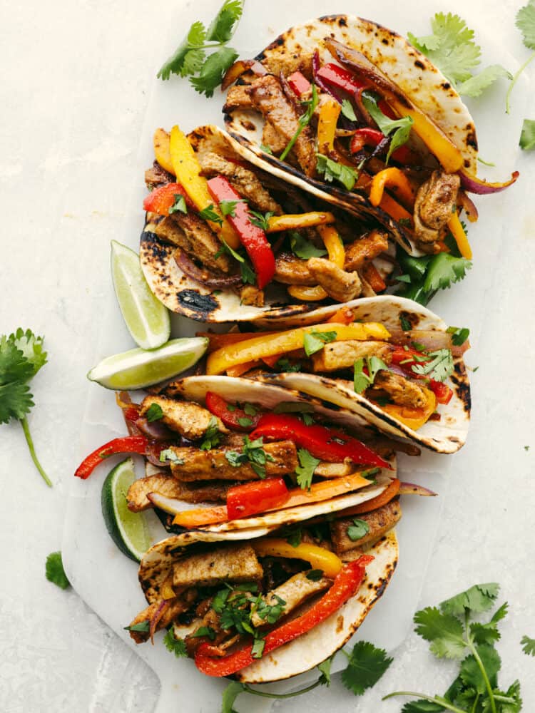 These pork fajitas are bursting with all your favorite Mexican flavors! The lean pork, fresh peppers and onions, fajita seasonings, and soft tortillas will have you coming back for more! Flour tortillas on a cutting board filled with seasoned sliced pork loin, bell peppers, and onions. Garnished with limes and cilantro.