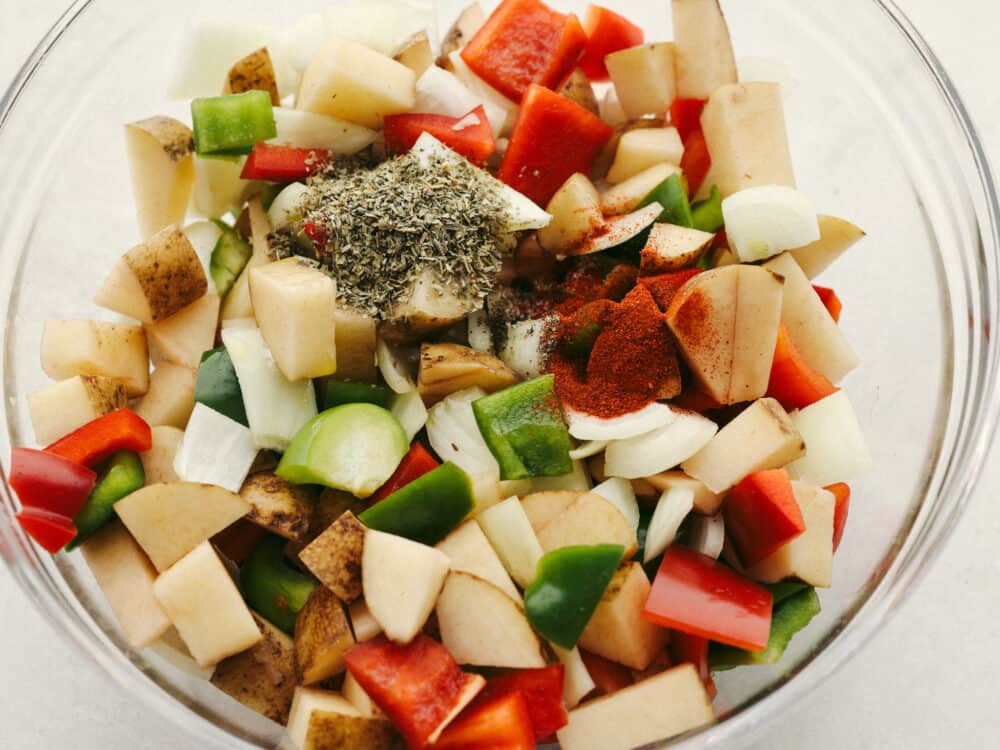 These breakfast potatoes make the best hearty and delicious breakfast! Filled with peppers, onions, and potatoes that are all coated with delicious homemade seasoning, this savory dish will become a family favorite! A clear, glass bowl with vegetables and seasonings ready to be mixed.