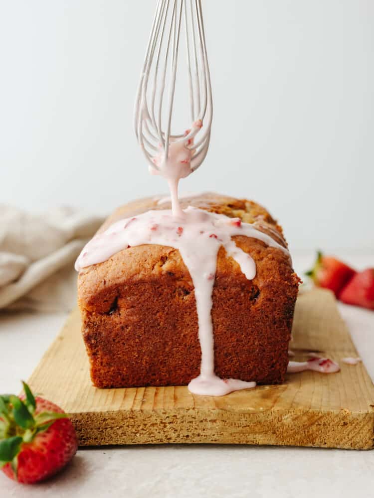 This strawberry quick bread is one of my all-time favorites! It is moist, packed with flavor, and drizzled with the most delightful strawberry glaze, which really brings all of the ingredients together. A whisk drizzling the strawberry glaze onto the loaf of quickbread.