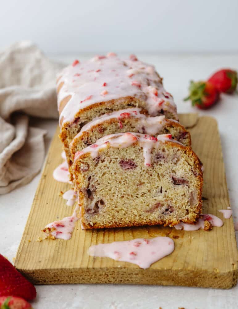 This strawberry quick bread is one of my all-time favorites! It is moist, packed with flavor, and drizzled with the most delightful strawberry glaze, which really brings all of the ingredients together. A loaf of strawberry quick bread sliced with a strawberry glaze sitting on a wooden cutting board.
