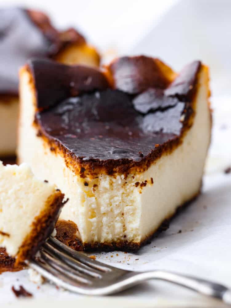 Try not to judge Basque cheesecake by its burnt outer layer because it's meant to be caramelized to perfection. You will be blown away by the rich and creamy Inside of this unique cheesecake. A close-up photo of a slice of Basque cheesecake. A piece of cheesecake on a fork lying next to the slice of cheesecake.