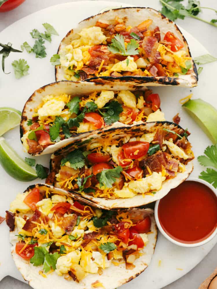 These delicious breakfast tacos are tortillas layered with potatoes, crispy bacon, soft scrambled eggs, and all the delicious toppings that you can think of! Filling and flavorful these tacos are the BEST way to start the day.ย Four breakfast tacos on a plate with a small bowl of ketchip and some lime wedges.