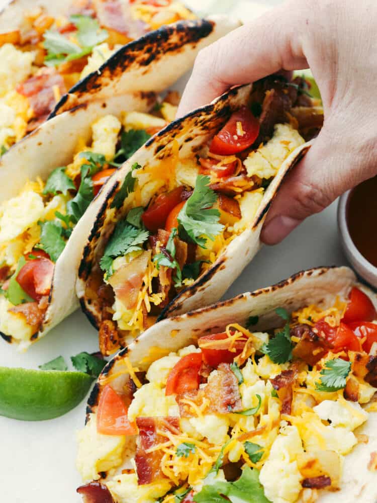 These delicious breakfast tacos are tortillas layered with potatoes, crispy bacon, soft scrambled eggs, and all the delicious toppings that you can think of! Filling and flavorful these tacos are the BEST way to start the day.ย A hand grabbing a fold breakfast taco.