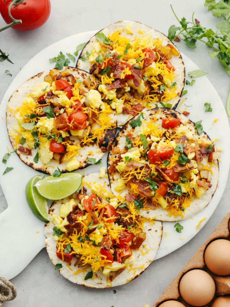 These delicious breakfast tacos are tortillas layered with potatoes, crispy bacon, soft scrambled eggs, and all the delicious toppings that you can think of! Filling and flavorful these tacos are the BEST way to start the day.ย 4 tortillas laying flat on a cutting board, each topped with breakfast taco filling.