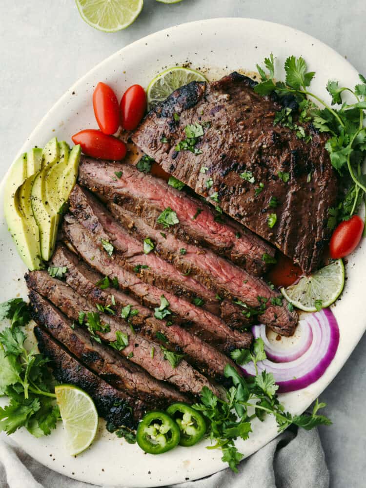 Enjoy this homemade carne asada recipe marinated in delicious Mexican flavors and spices. This juicy and tender flank steak is grilled to perfection for you to devour on its own or in any of your favorite Mexican dishes. Top view of sliced carne asada on a platter. Garnished with limes, cilantro, sliced avocado, tomatoes, red onion, and sliced japepeno.