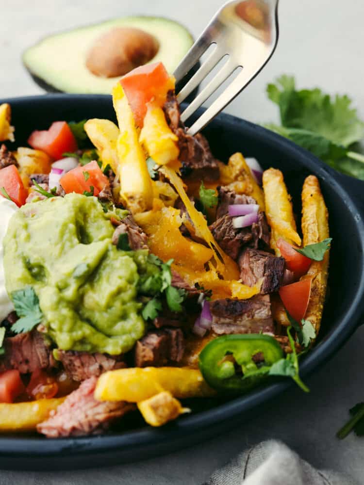 Skip the takeout from your favorite Mexican restaurant and try making carne asada fries at home instead! They're smothered in cheddar cheese, guacamole, jalapenos, and juicy carne asada for a flavor combination you won't be able to get enough of. Close-up of carne asada fries being stuck with a fork.