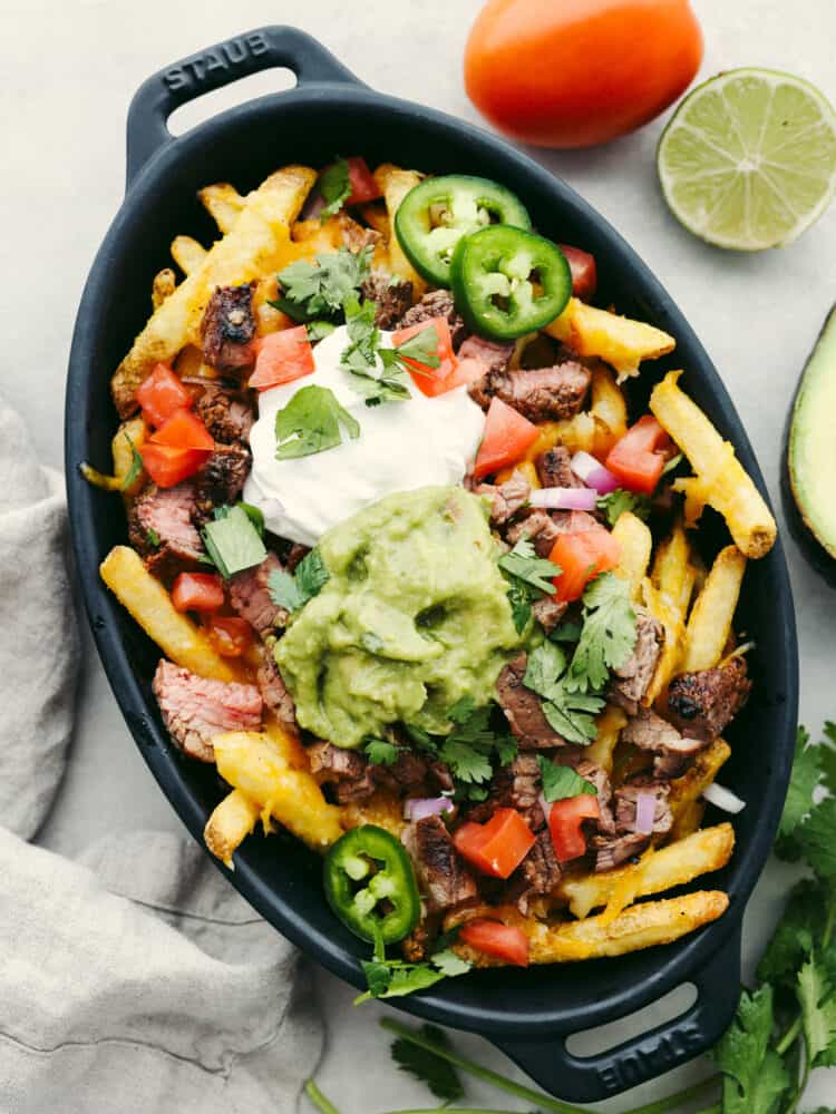 Skip the takeout from your favorite Mexican restaurant and try making carne asada fries at home instead! They're smothered in cheddar cheese, guacamole, jalapenos, and juicy carne asada for a flavor combination you won't be able to get enough of. Top-down view of carne asada fries in a black serving dish.