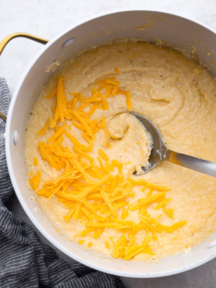 Creamy, cheesy, and comforting grits are a classic Southern dish. Slow-cooked cornmeal creates a deliciously smooth texture with a rich cheesy flavor. Top view of a large pot of cheesy grits with a large metal spoon laying in the grits. Shredded cheddar cheese garnished on top with a gray kitchen bowl on the side of the pot.