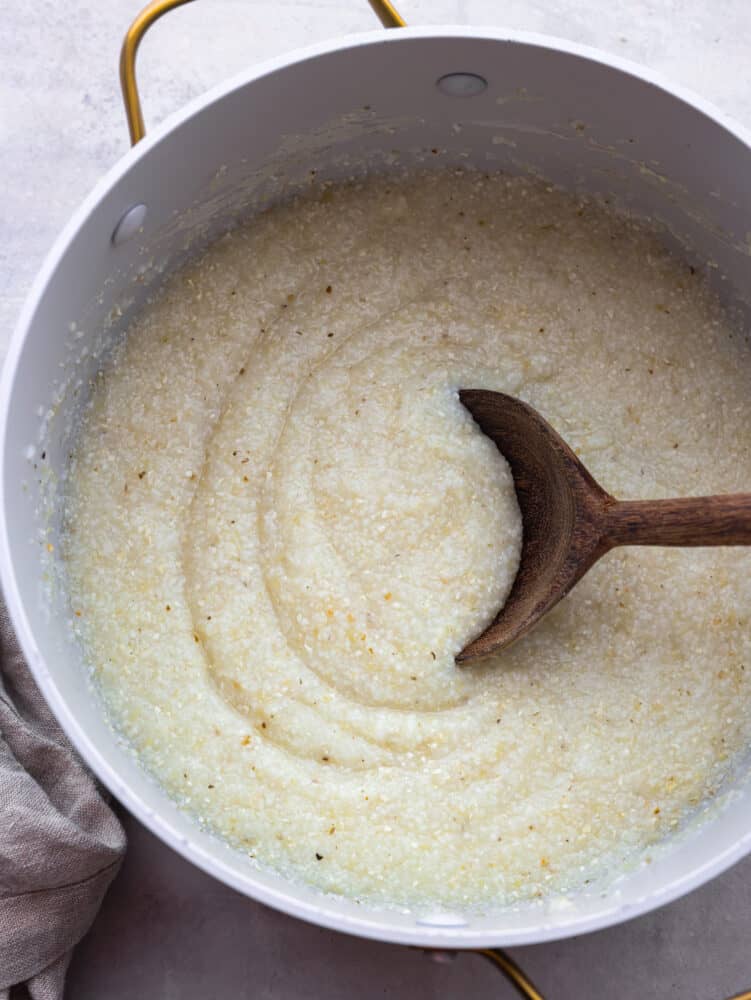 Southern grits are the perfect breakfast (or lunch or dinner!) option. They are the creamiest and most delicious addition to your morning meal, and everyone in your family will be able to make these. They are that easy! A pot of cooked grits with a wooden spoon.