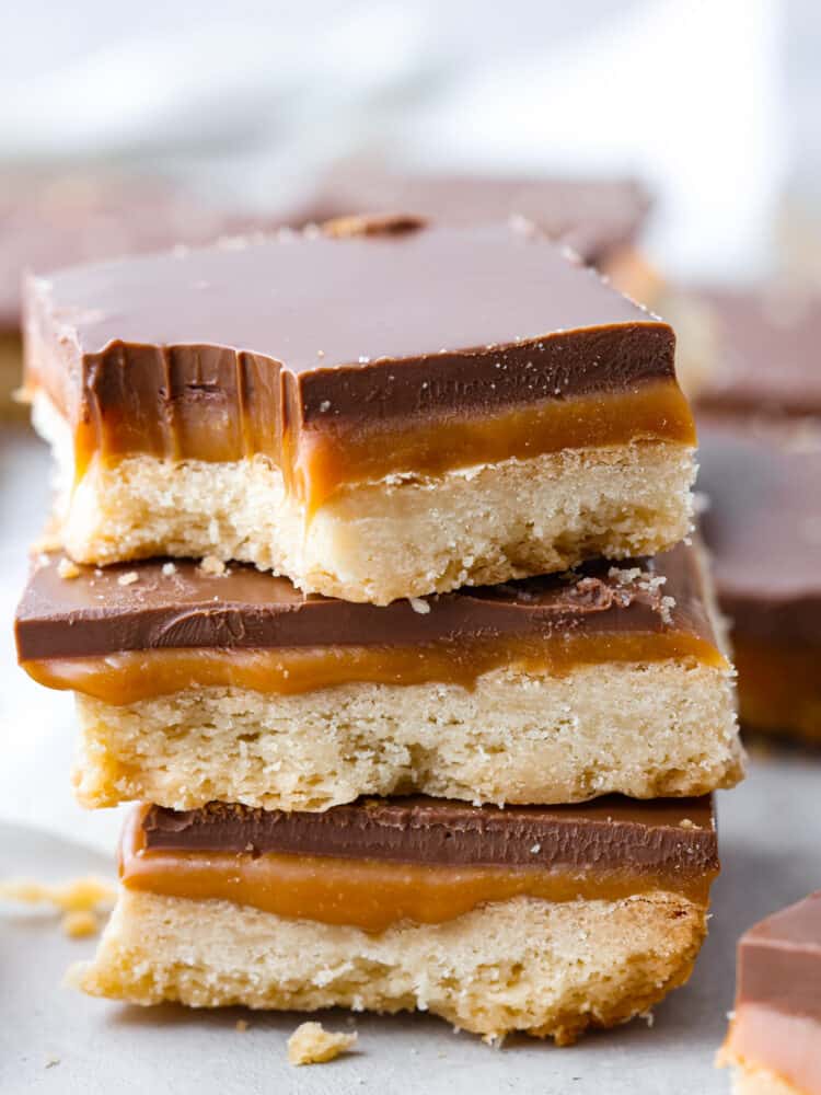 These taste exactly like a Twix bar with a delicious shortbread crust, caramel center, and a chocolate layer on top. The best part about them is that you can make them right at home! A stack of homemade Twix bars, the top one has a bite taken out of it.