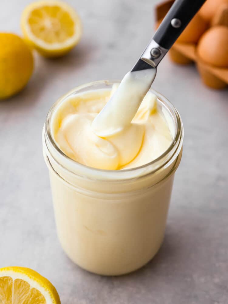 Easy homemade mayonnaise is fresh and convenient to make right at home. With just a few ingredients, you can make this delicious and versatile condiment to use on sandwiches or in dips and sauces! Close up photo of creamy mayonnaise in a mason jar with a knife lifting out mayonnaise. Lemons and eggs in the background.