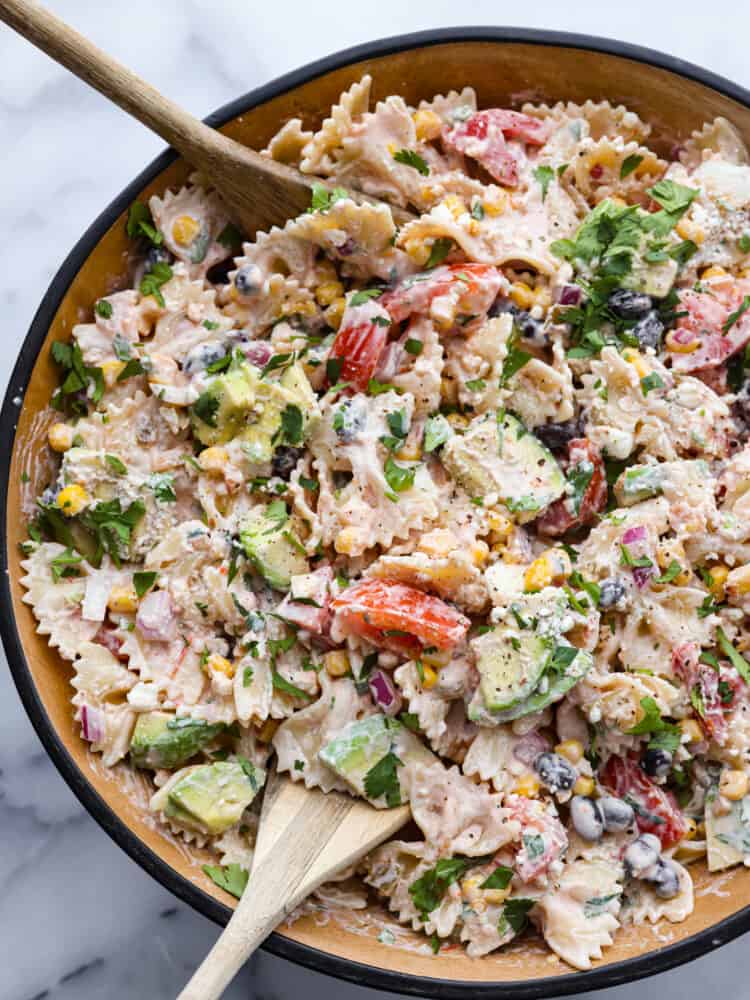 Mexican Pasta Salad is an unbelievably easy and flavorful pasta salad that is filled with colorful ingredients like black beans, tomatoes, and corn.ย With a creamy dressing and spices from onion, cilantro, and cumin, this delicious pasta salad will be the hit of your summer barbecues for sure! A bowl of Mexican pasta salad with a wooden spoon.