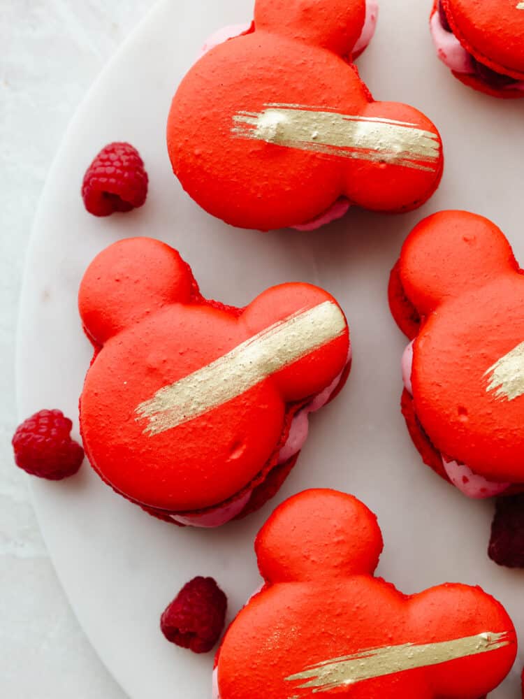Inspired by Jolly Holiday Bakery Cafe in Disneyland, these raspberry rose Mickey macarons are a beautiful and delicious dessert. A light and airy shell paired with a creamy, raspberry mousse filling makes them absolutely irresistible. Top-down view of Mickey Mouse-shaped macarons on a white plate.