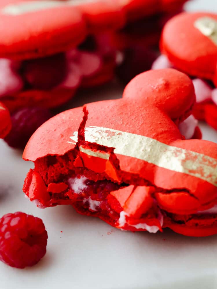 Inspired by Jolly Holiday Bakery Cafe in Disneyland, these raspberry rose Mickey macarons are a beautiful and delicious dessert. A light and airy shell paired with a creamy, raspberry mousse filling makes them absolutely irresistible. Closeup of a cookie with a bite taken out of it.