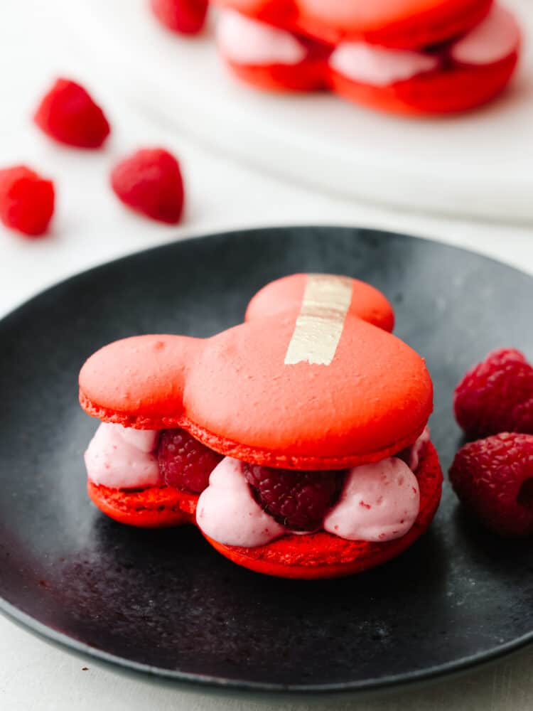 Inspired by Jolly Holiday Bakery Cafe in Disneyland, these raspberry rose Mickey macarons are a beautiful and delicious dessert. A light and airy shell paired with a creamy, raspberry mousse filling makes them absolutely irresistible. Closeup of a mickey macaron on a black plate, raspberry filling can be seen.
