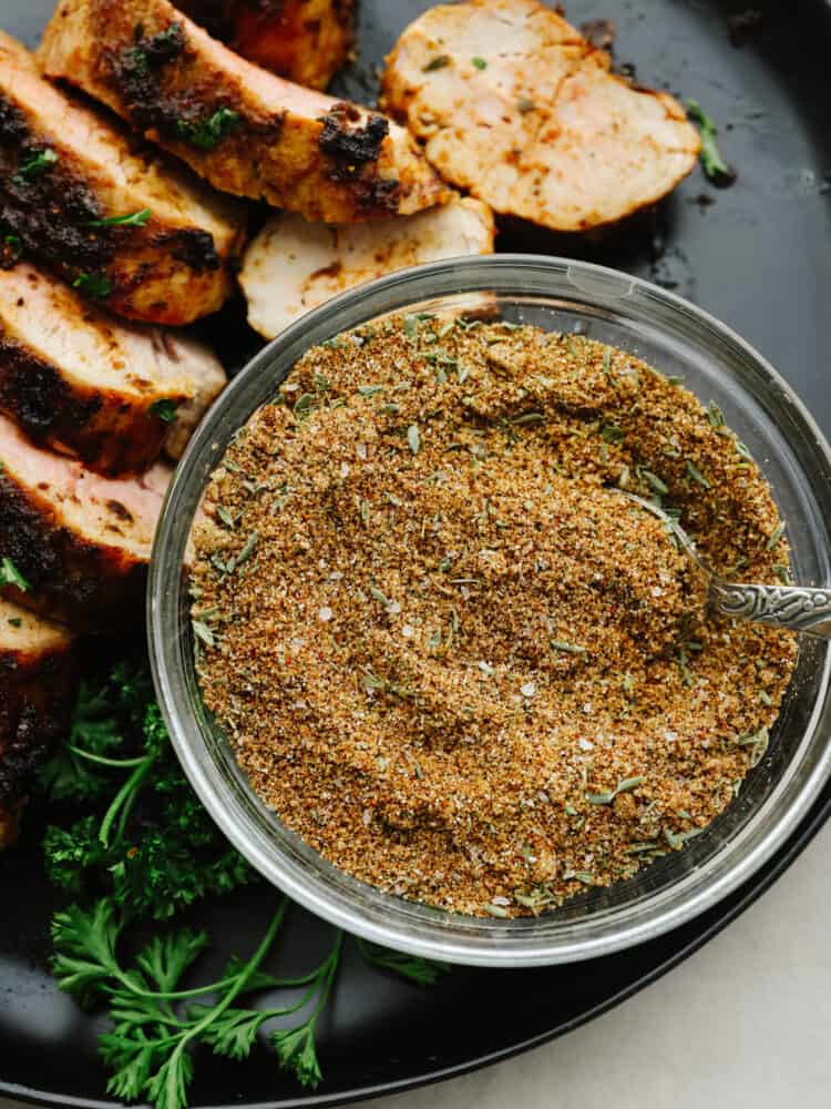 This pork seasoning is the perfect bled of delicious spices. It only takes about 5 minutes to whip up and will elevate your meals for months to come! Pork seasoning in a bowl with a spoon on a platter with sliced, cooked pork.