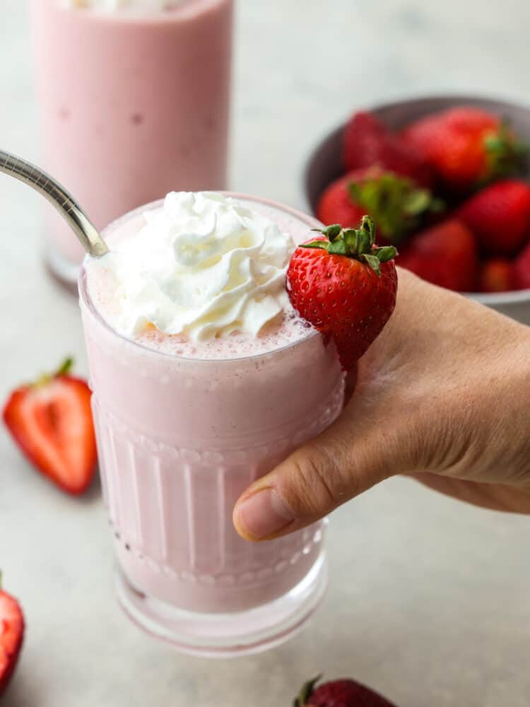 This strawberry milkshake is thick, creamy, and everything that you would hope a milkshake would be! Made with delicious strawberries, this classic dessert is sure to be loved by all! A hand holding a glass filled with strawberry milkshake. There is a silver straw in the cup and a fresh strawberry on the side of the glass.