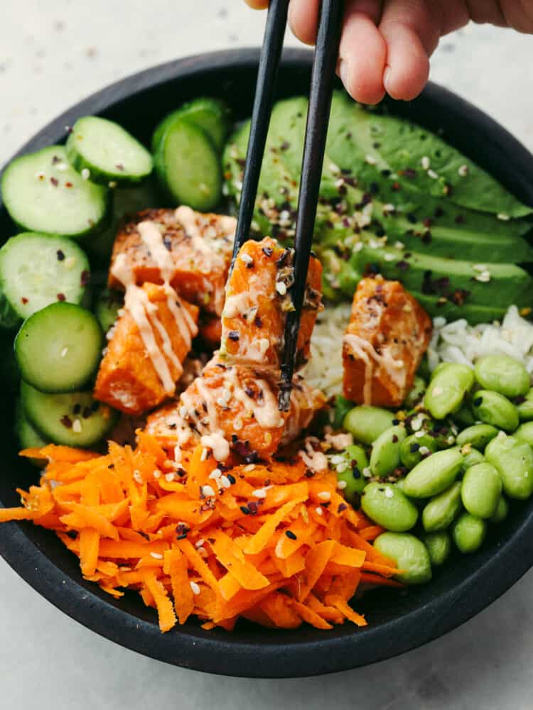 Perfect air fryer salmon nestled in a bowl of Asian-inspired ingredients. The fluffy rice topped with fresh vegetables and a sriracha mayonnaise creates this delicious salmon rice bowl that will become your new favorite! Top view of the assembled salmon rice bowl with chopsticks lifting out the salmon from the bowl.