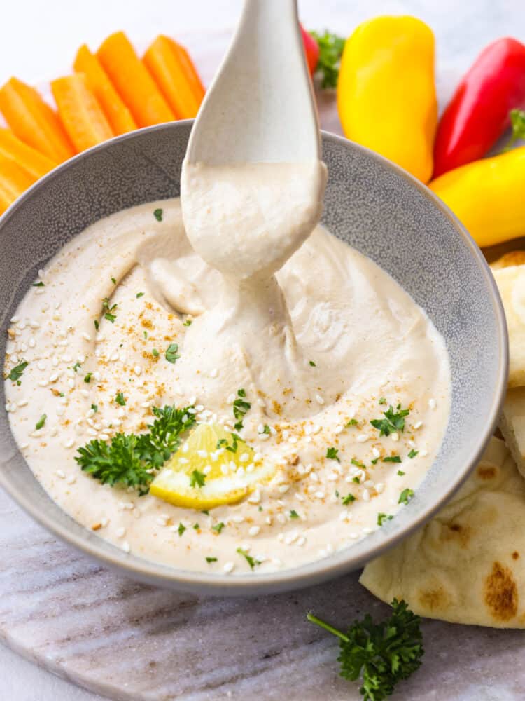 Get ready for the best tahini sauce that you have ever tasted! This makes any falafel, vegetable or salad taste better and it takes less than 5 minutes to put it all together! A bowl of tahini sauce on a platter with veggies. A plastic seerving spoon is scooping out some sauce.