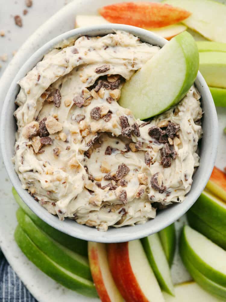 Toffee apple dip is a family favorite! A sweet dip complete with heath bar candy bits and cream cheese mixed with brown sugar, this dip is so delicious you won't be able to stop eating it! An apple slice in a bowl of toffee apple dip.