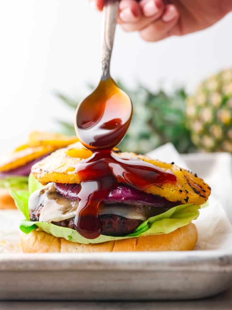 These tangy and zesty burgers are topped with teriyaki sauce, pineapple, and all the delicious hamburger fixings that you could want. It's dripping with flavor and will soon become a family favorite! Teriyaki sauce being drizzled on the inside hamburger ingredients with a silver spoon.