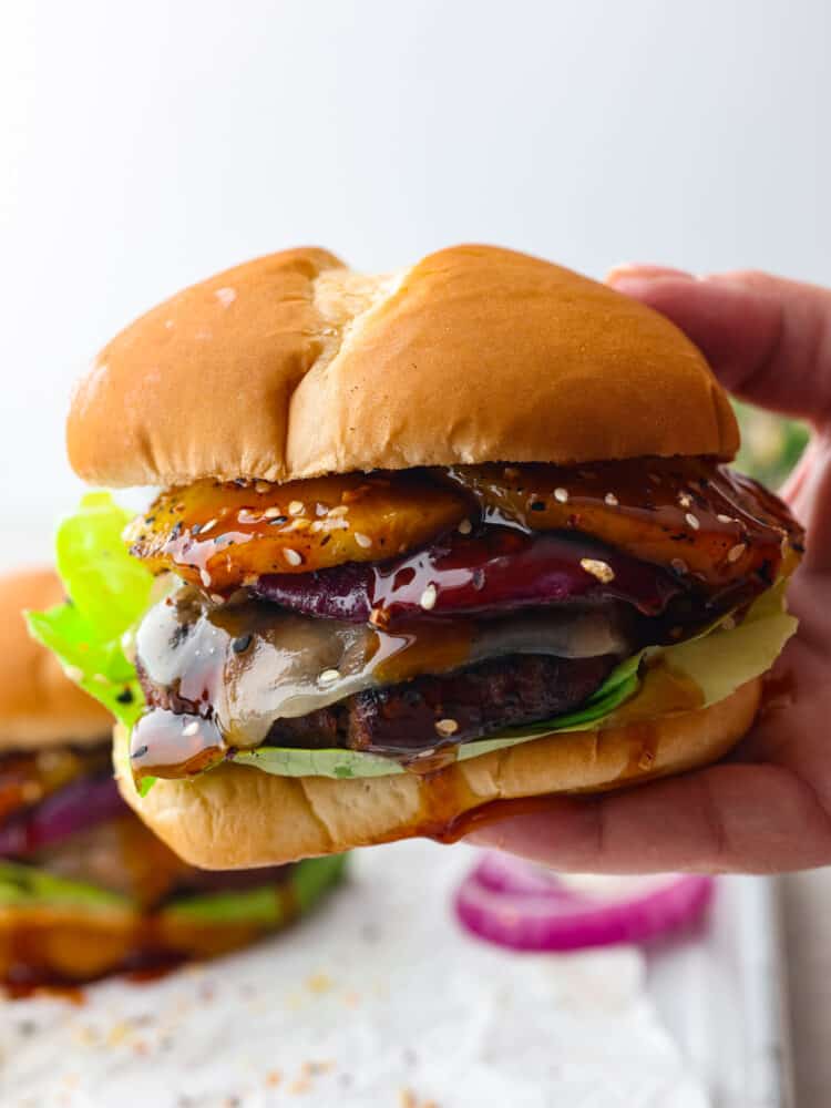 These tangy and zesty burgers are topped with teriyaki sauce, pineapple, and all the delicious hamburger fixings that you could want. It's dripping with flavor and will soon become a family favorite! A hand holding a teriyaki burger that's ready to eat.