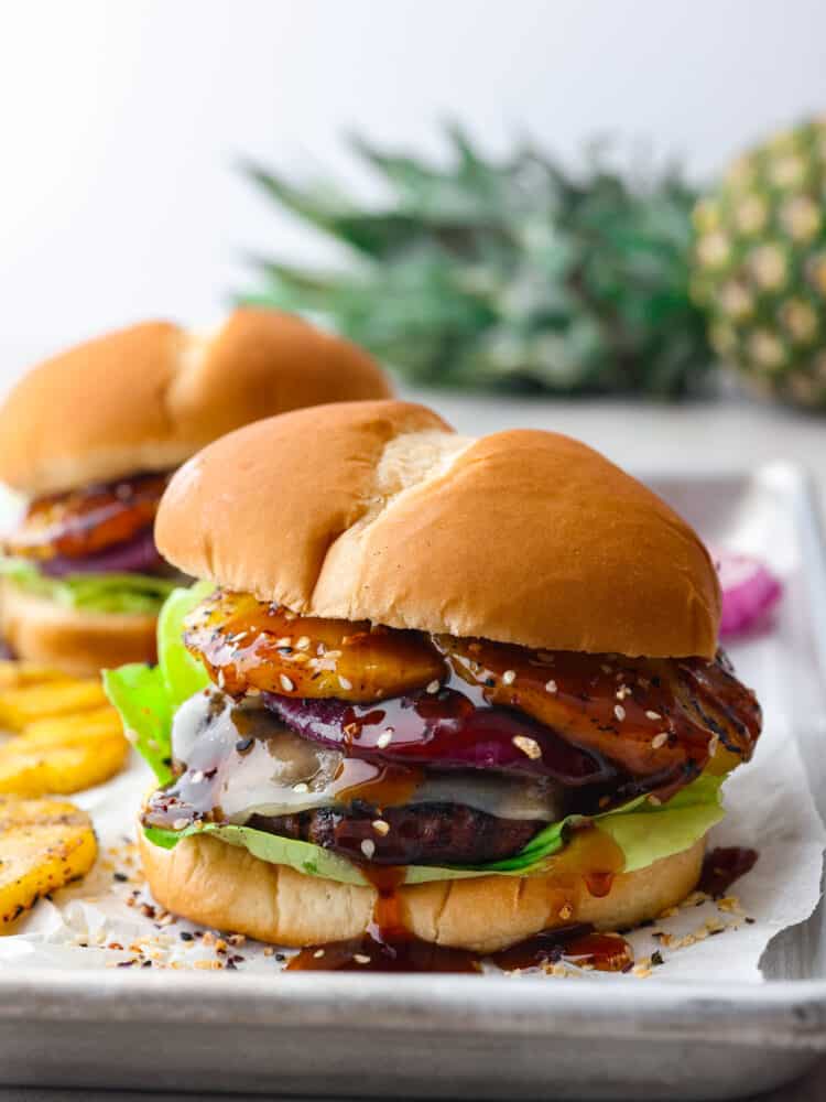 These tangy and zesty burgers are topped with teriyaki sauce, pineapple, and all the delicious hamburger fixings that you could want. It's dripping with flavor and will soon become a family favorite! The side view of a teriyaki burger on a baking sheet with parchment paper.