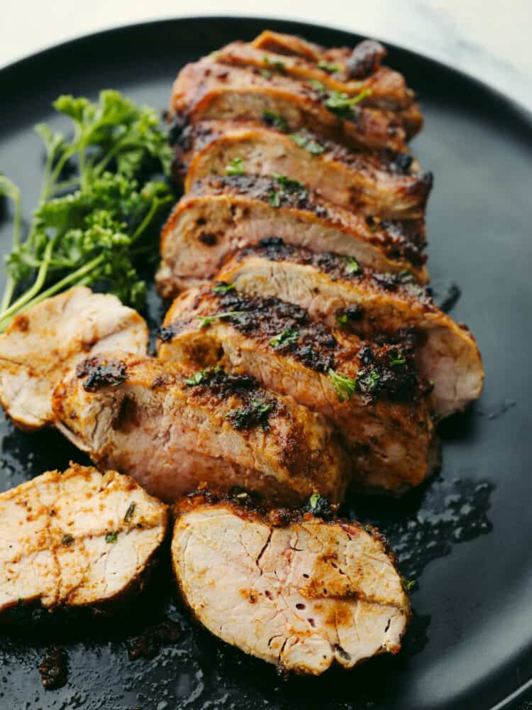 Air fryer pork tenderloin is the ultimate weeknight dinner hero! It's quick, juicy, and packed with flavor! It cooks up perfectly with minimal effort, thanks to a simple marinade and the magic of the air fryer. Close up photo of cooked sliced tenderloin on a black plate. Garnished with a sprig of parsley on the side.