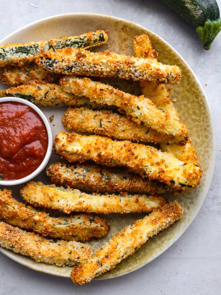 Air fryer zucchini fries are a low-carb, keto-friendly snack that's ready in minutes! Crispy and cheesy on the outside, tender on the inside, this is the best way to use up your leftover summer zucchini. Air-fried zucchini fries served with marinara sauce on a gray plate.