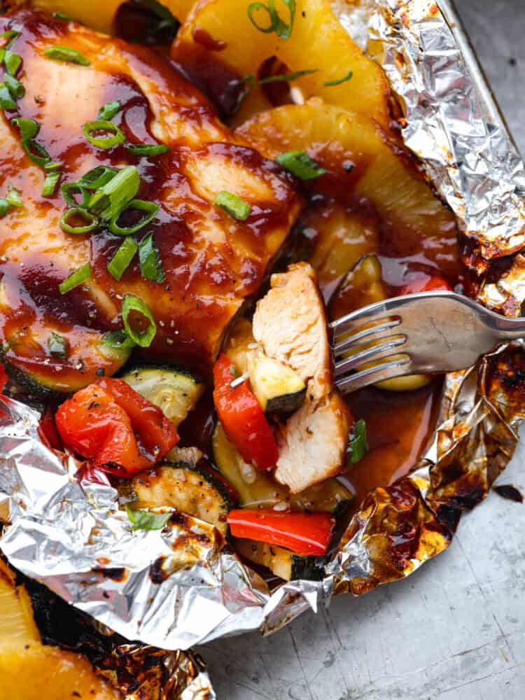 Grilled Hawaiian barbecue chicken that is made in foil has the most amazing sweet and tangy pineapple barbecue sauce! It grills to perfection with fresh pineapple and delicious summer veggies! A close up of a fork cutting into the grilled Hawaiian barbecue chicken thats in a foil packet.