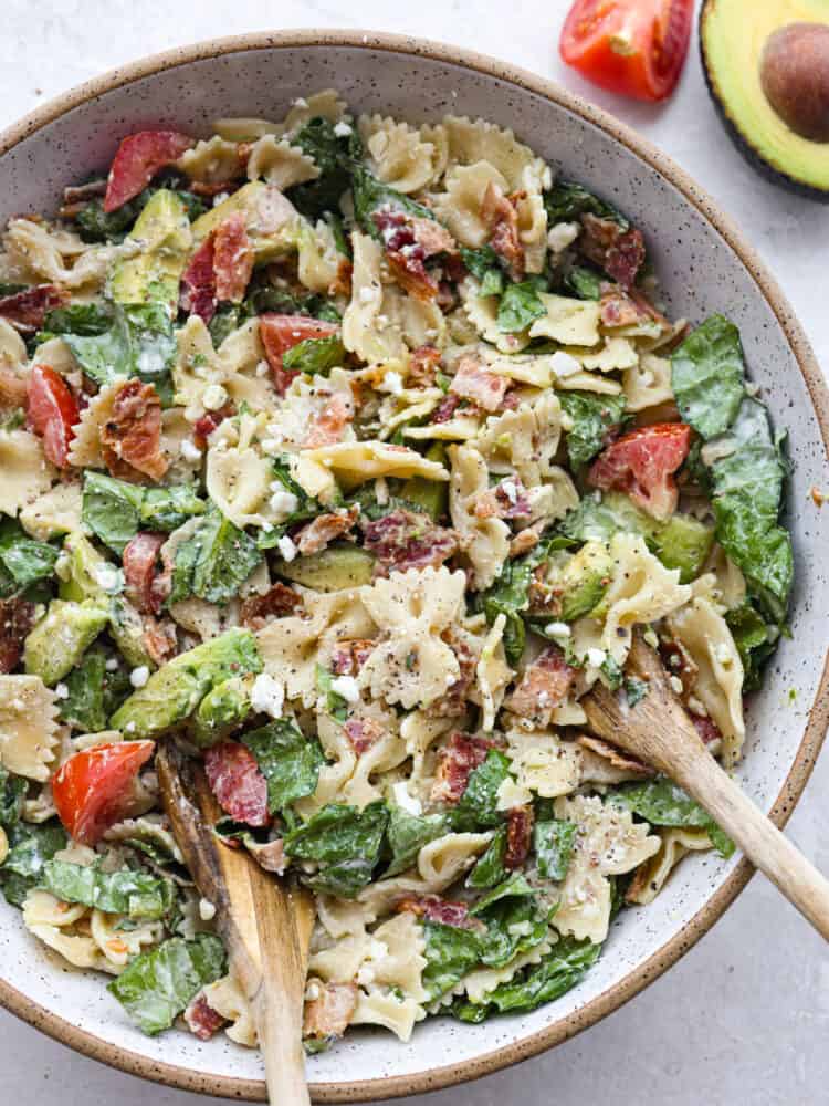 BLT Avocado Pasta Salad is fresh and crisp romaine lettuce tossed in bacon, tomato, pasta, and avocado. Topped with a creamy dressing and sprinkled with feta cheese this is one amazing salad! A bowl filled with pasta salad, topped with feta and being tossed with two wooden spoons.