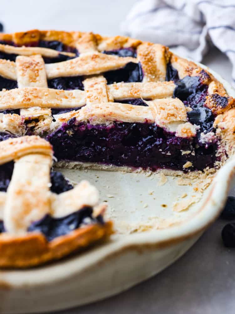 This classic blueberry pie is a fresh and delicious dessert that's made for summertime! Filled with blueberries and topped with the most delicious and beautiful lattice crust, this pie is sure to impress any guest. The side view of a blueberry lattice pie in a pie pan with a slice taken out.
