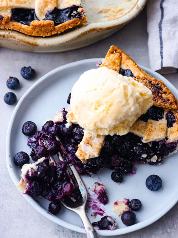 This classic blueberry pie is a fresh and delicious dessert that's made for summertime! Filled with blueberries and topped with the most delicious and beautiful lattice crust, this pie is sure to impress any guest. A slice of blueberry pie with a scoop of vanilla ice cream on a periwinkle colored plate.