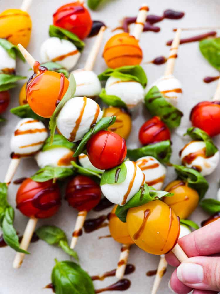 Caprese skewers are a 4-ingredient appetizer that is so simple but guaranteed to impress! Made with cherry tomatoes, mozzarella, fresh basil, and an easy balsamic reduction, they imitate classic Caprese salad, only made portable on a skewer! Closeup of a Caprese skewer being held.