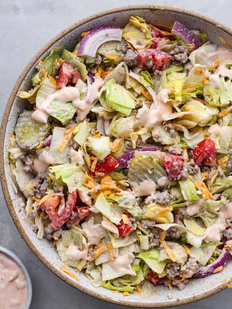 Cheeseburger salad is a delicious bowl of all your favorite burger ingredients, just without the bun. This healthier low-carb version of a juicy cheeseburger is loaded with seasoned beef, cheddar cheese, tomatoes, onion, lettuce, and pickles, then tossed in a delicious dressing. Top view of the cheeseburger salad in a large tan bowl. The salad is tossed with a thousand island dressing with a small bowl of the dressing on the side.