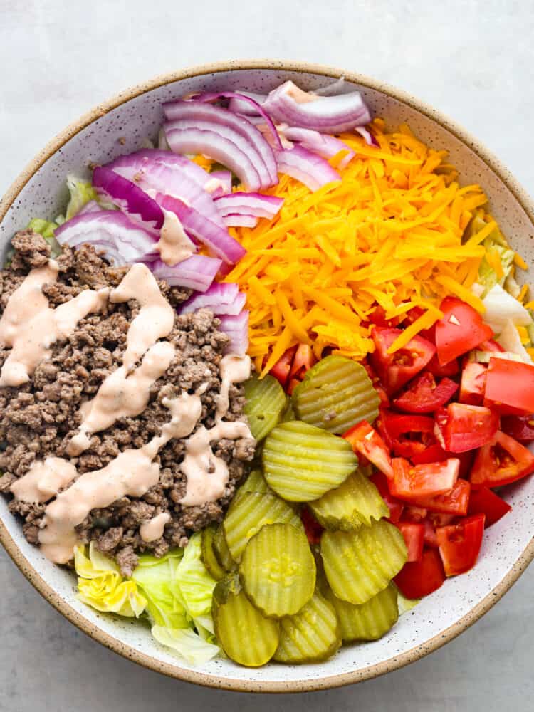 Cheeseburger salad is a delicious bowl of all your favorite burger ingredients, just without the bun. This healthier low-carb version of a juicy cheeseburger is loaded with seasoned beef, cheddar cheese, tomatoes, onion, lettuce, and pickles, then tossed in a delicious dressing. A top view of salad ingredients in a large bowl before they are tossed together. The thousand island dressing is drizzled over the cooked meat.