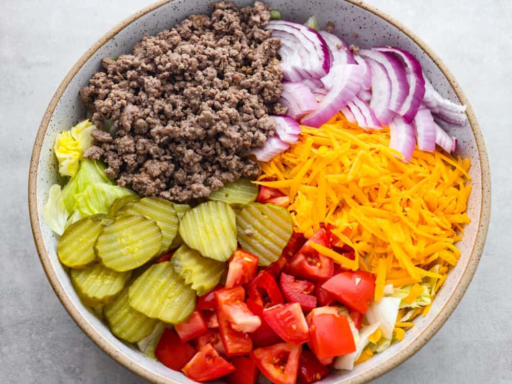 Cheeseburger salad is a delicious bowl of all your favorite burger ingredients, just without the bun. This healthier low-carb version of a juicy cheeseburger is loaded with seasoned beef, cheddar cheese, tomatoes, onion, lettuce, and pickles, then tossed in a delicious dressing. A top view of the salad ingredients separately sectioned in a large bowl.