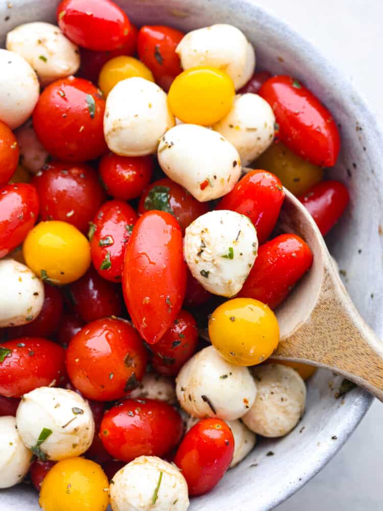 This cherry tomato salad is made with flavorful herbs, delicious cherry tomatoes, and fresh mozzarella! Coated in a delicious lemon basil dressing, this salad will soon be your new favorite! Cherry tomato salad in a bowl being served with a wooded spoon.