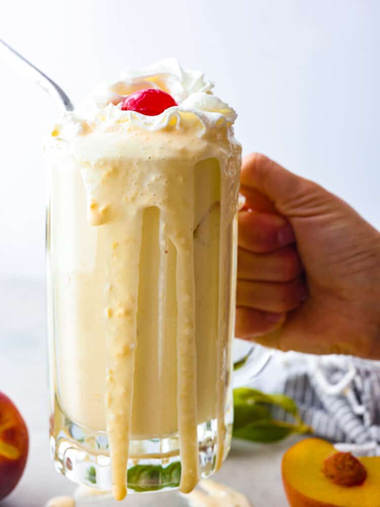 Chick-fil-A's peach shakes are the best summer treat! Now you can enjoy this delicious seasonal drink all year long with this quick and easy copycat recipe. It's too creamy and indulgent to pass up! Holding a peach milkshake in a glass mug, with the milkshake spilling down the sides.